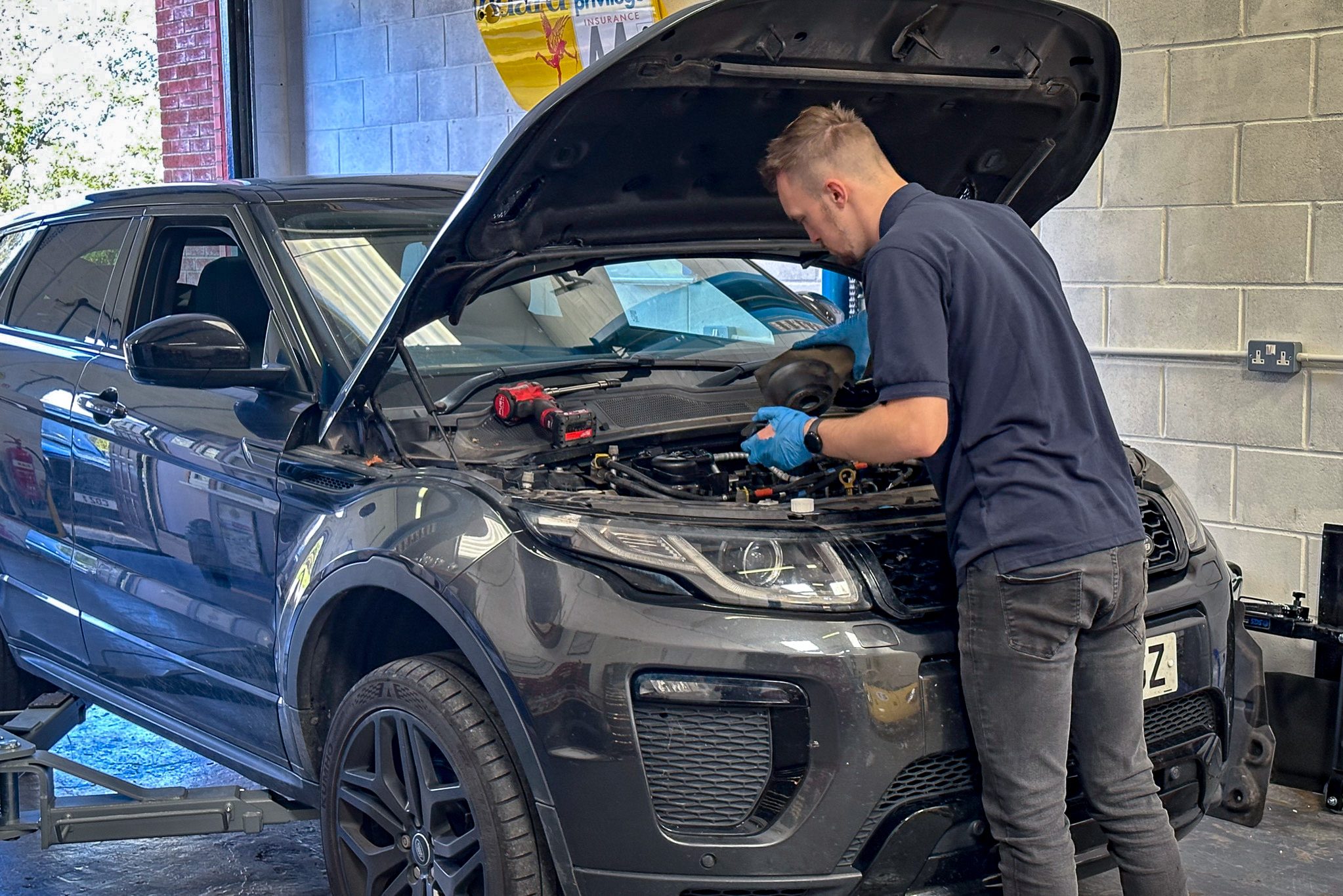 A skilled mechanic from Coastal Motorsport a Garage North Walsham is engaged in servicing a Range Rover. He stands with focus and precision as he works under the raised bonnet, with various tools laid out beside him, indicative of the thorough maintenance being performed. The vehicle's sleek black exterior is contrasted against the bright and orderly garage interior, reflecting the high standards of vehicle care and expertise offered.