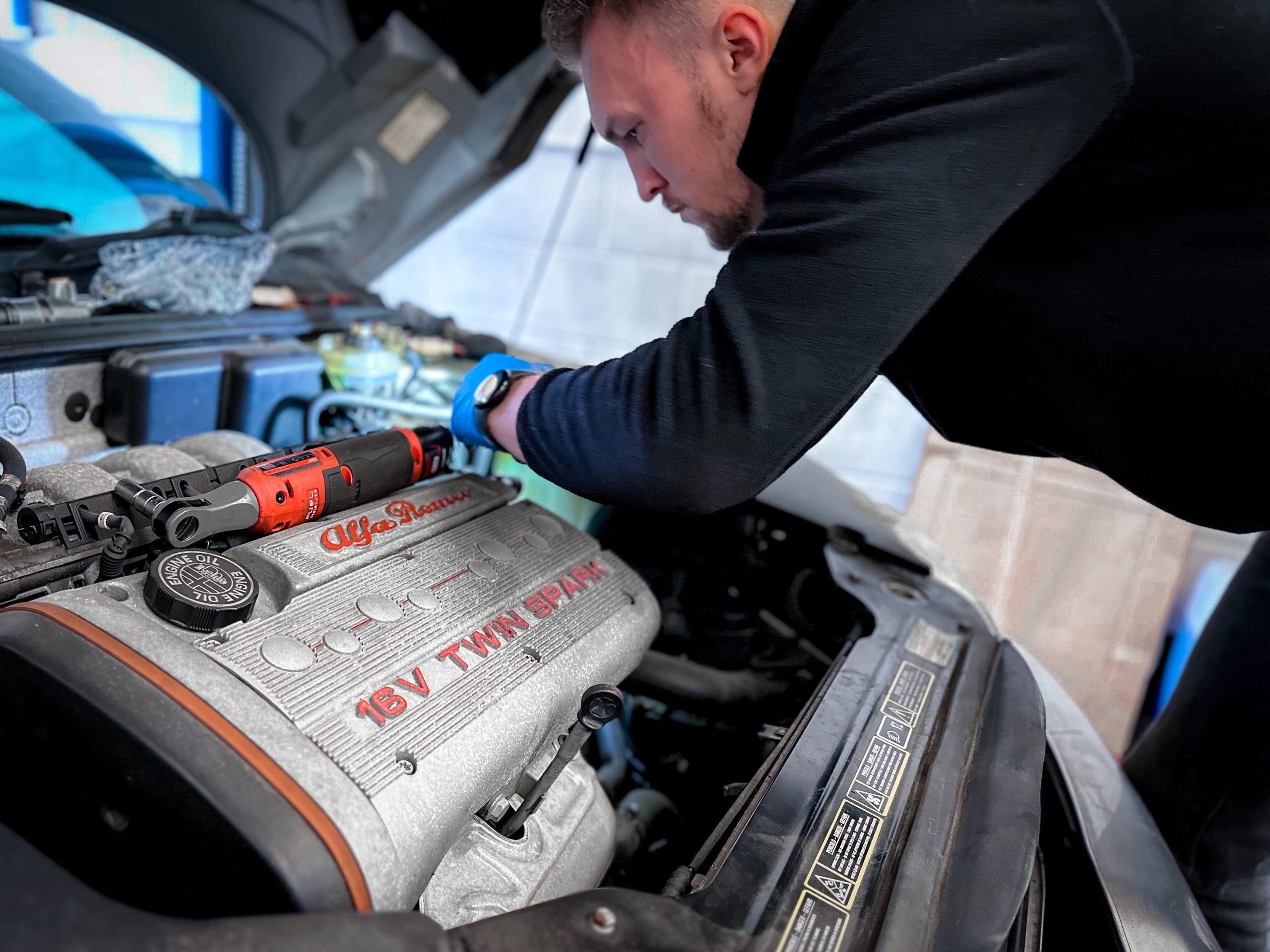 A mechanic is servicing a car in North Walsham, focusing intently on the engine compartment. They are wearing a black long-sleeve top and are in the process of working on an Alfa Romeo 16V Twin Spark engine. A black and orange cordless drill, along with various tools, is laid out on the engine cover, indicating an ongoing maintenance task. The bonnet is open, revealing the intricate components of the engine bay.