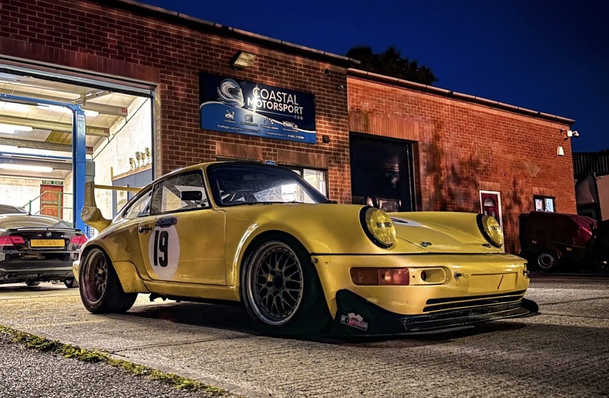A vintage yellow Porsche with the number 19 on its side is prominently positioned outside Coastal Motorsport garage north walsham, under the dusky evening sky in North Walsham. The iconic car, showcasing a racing heritage, is framed by the open garage doors revealing a well-lit and professional workspace within, and a variety of other vehicles being serviced, highlighting the garage's expertise with high-performance and classic cars. The scene exudes a blend of automotive excellence and the inviting atmosphere of the establishment.
