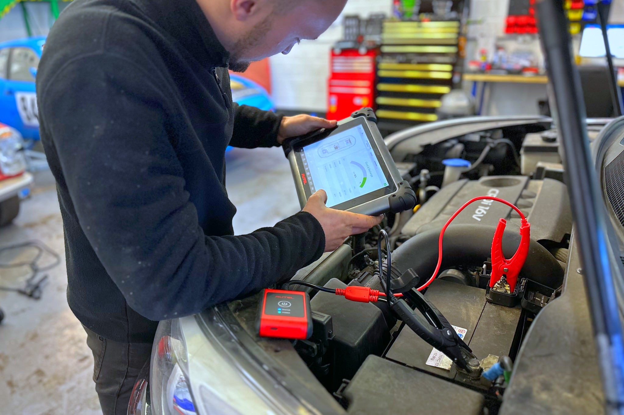 A mechanic in a black fleece is conducting a diagnostic test on a vehicle at a garage North Walsham. The technician is focused on a handheld diagnostic tool displaying vehicle stats. The car battery has red and black jumper cables attached, and the environment suggests an in-depth servicing session. Tools and equipment are visible in the background, indicating a professional automotive workshop setting.