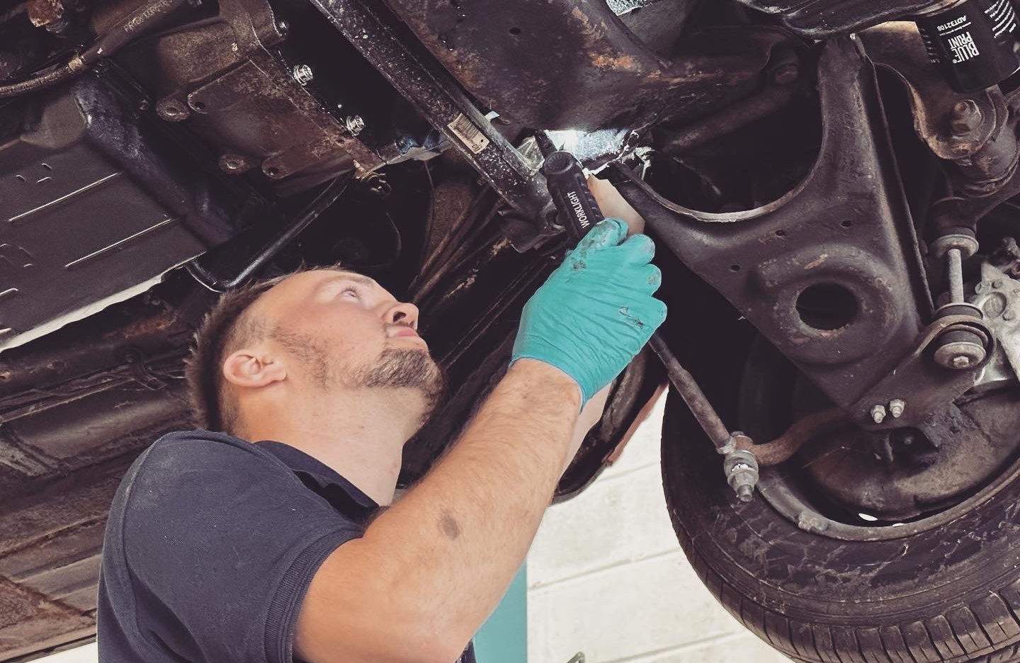 A mechanic is attentively working underneath a vehicle, suspended on a lift, at Coastal Motorsport a garage in North Walsham. Wearing dark work attire paired with bright teal gloves, he's engaged in adjusting or inspecting the vehicle's underbody, possibly focusing on the suspension system. The image showcases an intricate view of the car’s undercarriage with various mechanical components visible, portraying the precision and expertise required in automotive maintenance.